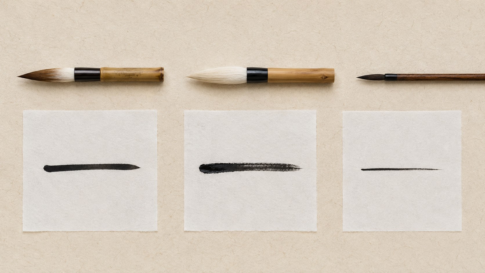 Three Japanese calligraphy brushes laid horizontally on a cream background, each with the same kanji 一 (ichi) brushstroke beneath it, showing how a mixed-hair, pure goat-hair, and small kofude brush each produce a different stroke character.