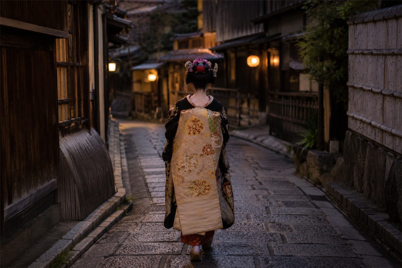 A Kyoto maiko seen from behind in a quiet stone alley, wearing an elaborate kimono with a long trailing obi and ornamental hairpins, walking toward the lantern light of an old teahouse district at dusk.