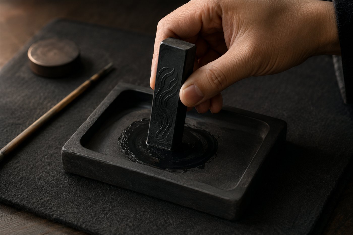 A hand holding an upright black sumi inkstick, grinding it in slow circles on a dark rectangular inkstone (suzuri), a pool of glossy fresh black ink gathered in the well, on a felt mat.