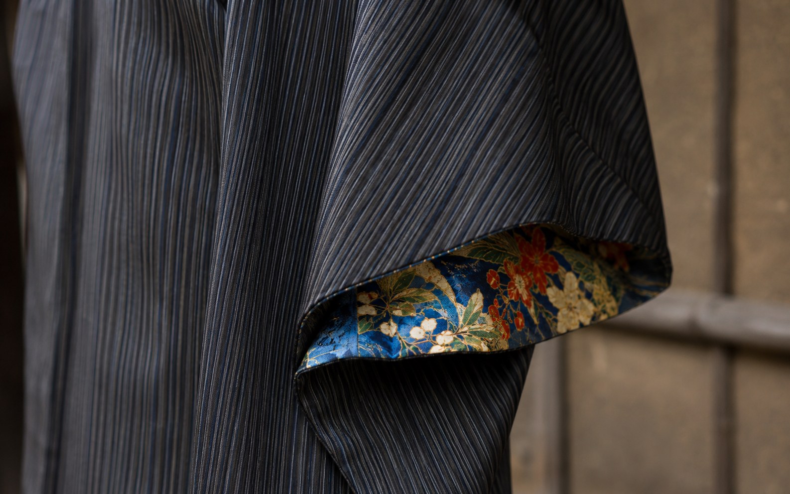 A close-up of an Edo-style kimono detail: a plain, finely vertical-striped indigo-and-grey outer fabric falling open just enough to reveal a flash of a brilliant, richly colored silk lining — hidden luxury glimpsed rather than displayed, the essence of iki.