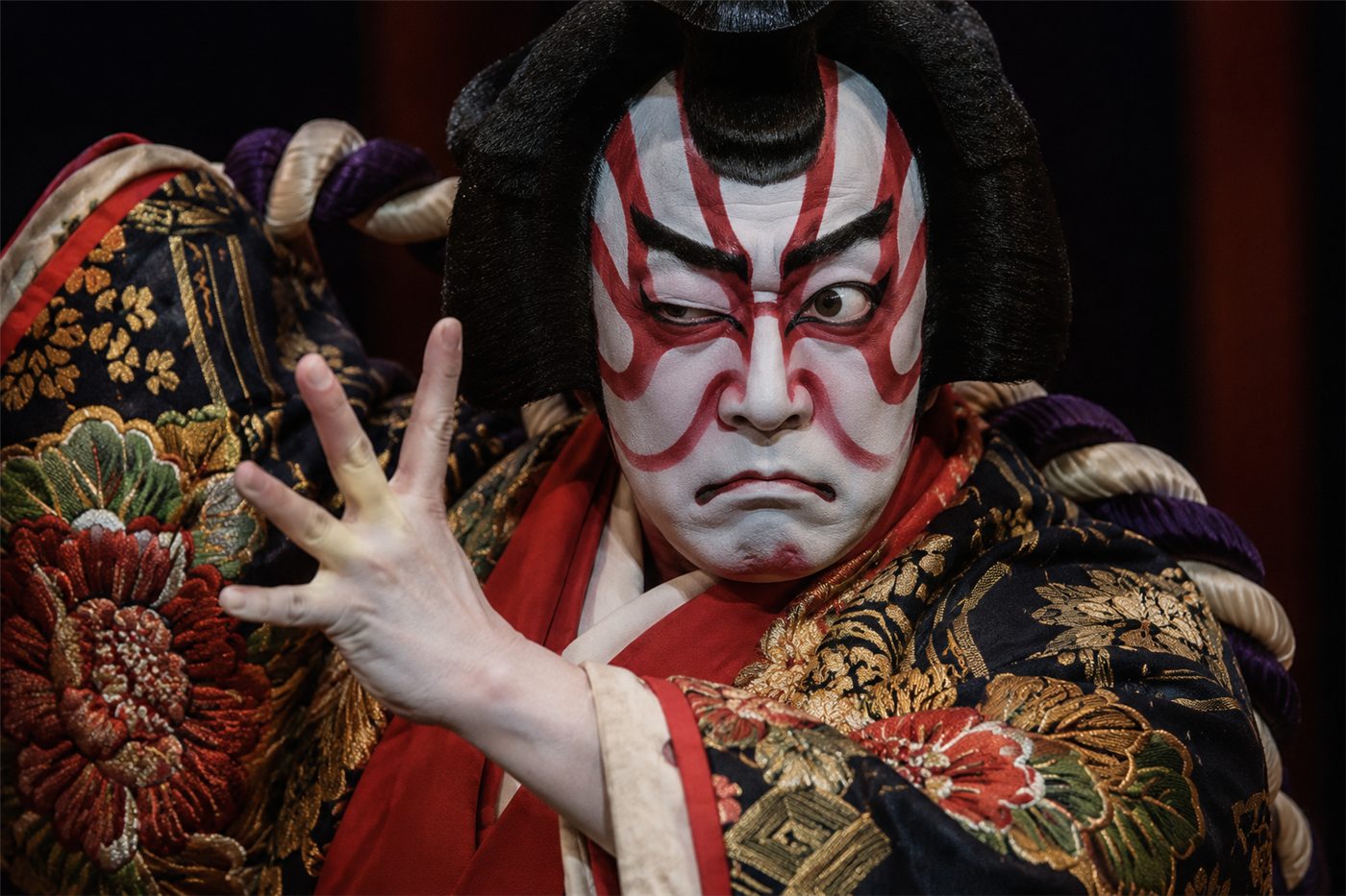 A kabuki actor in bold red-and-white kumadori makeup, striking a dramatic frozen mie pose with one eye crossed, in an ornate patterned costume against a dark stage.