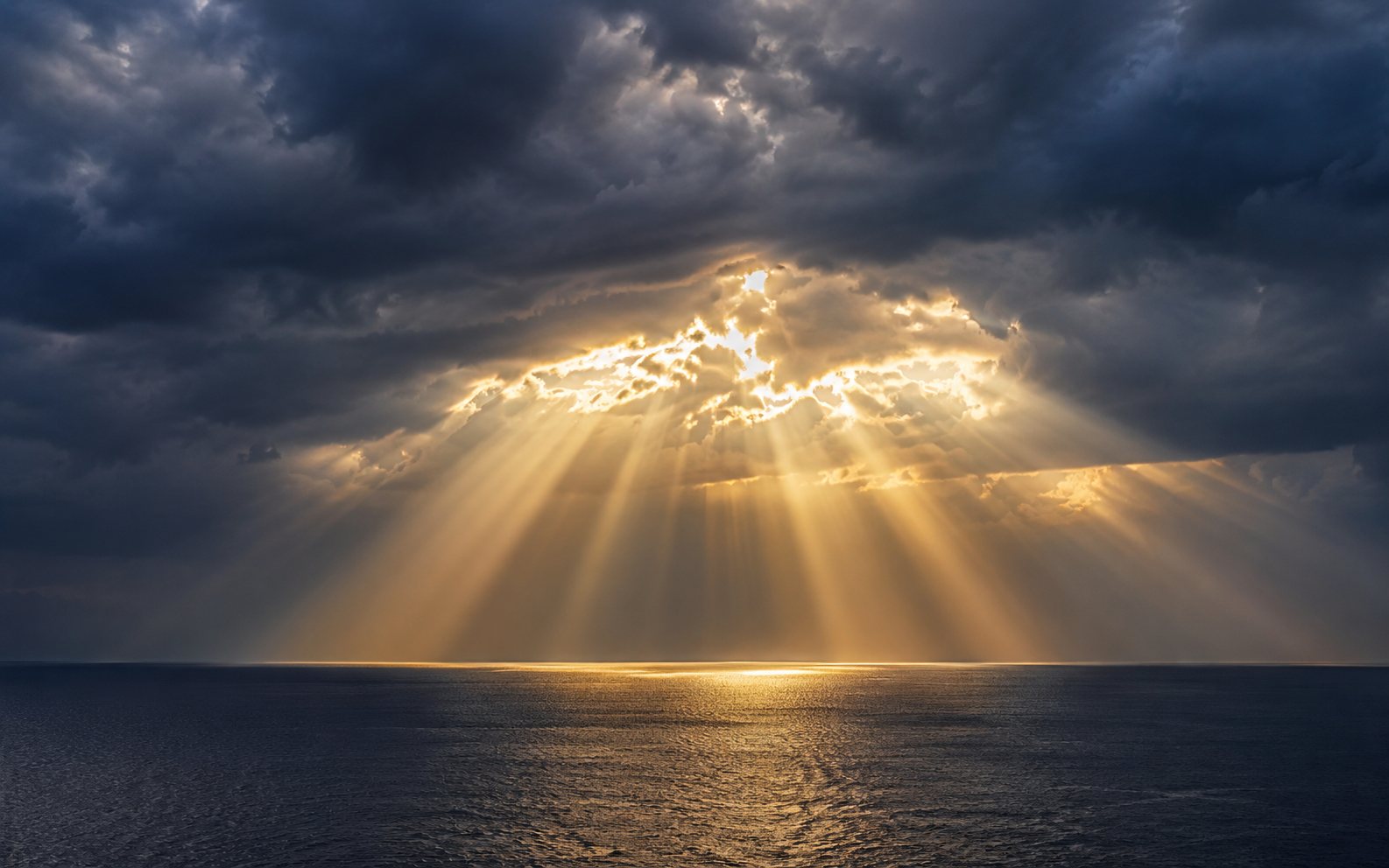 Brilliant rays of sunlight breaking through a gap in dark clouds and fanning down toward the sea — the physical radiance the kanji 光 names.