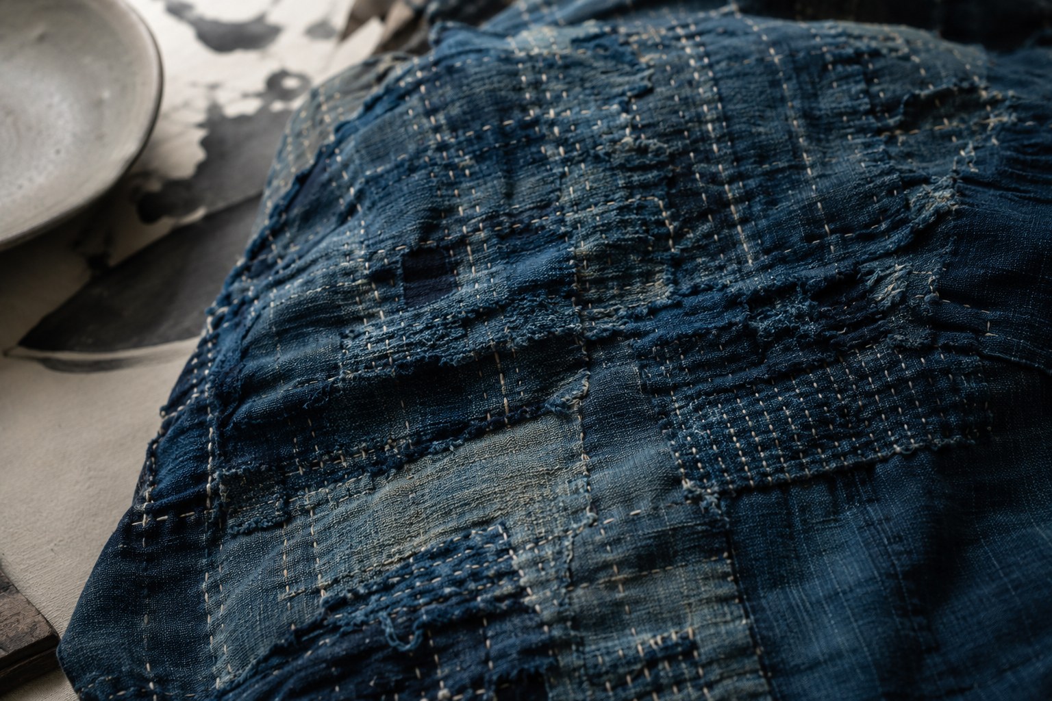 A close-up of indigo boro textile — old cloth mended with many layers of sashiko stitching and patches, the worth of the worn fabric honored by repair rather than replacement.