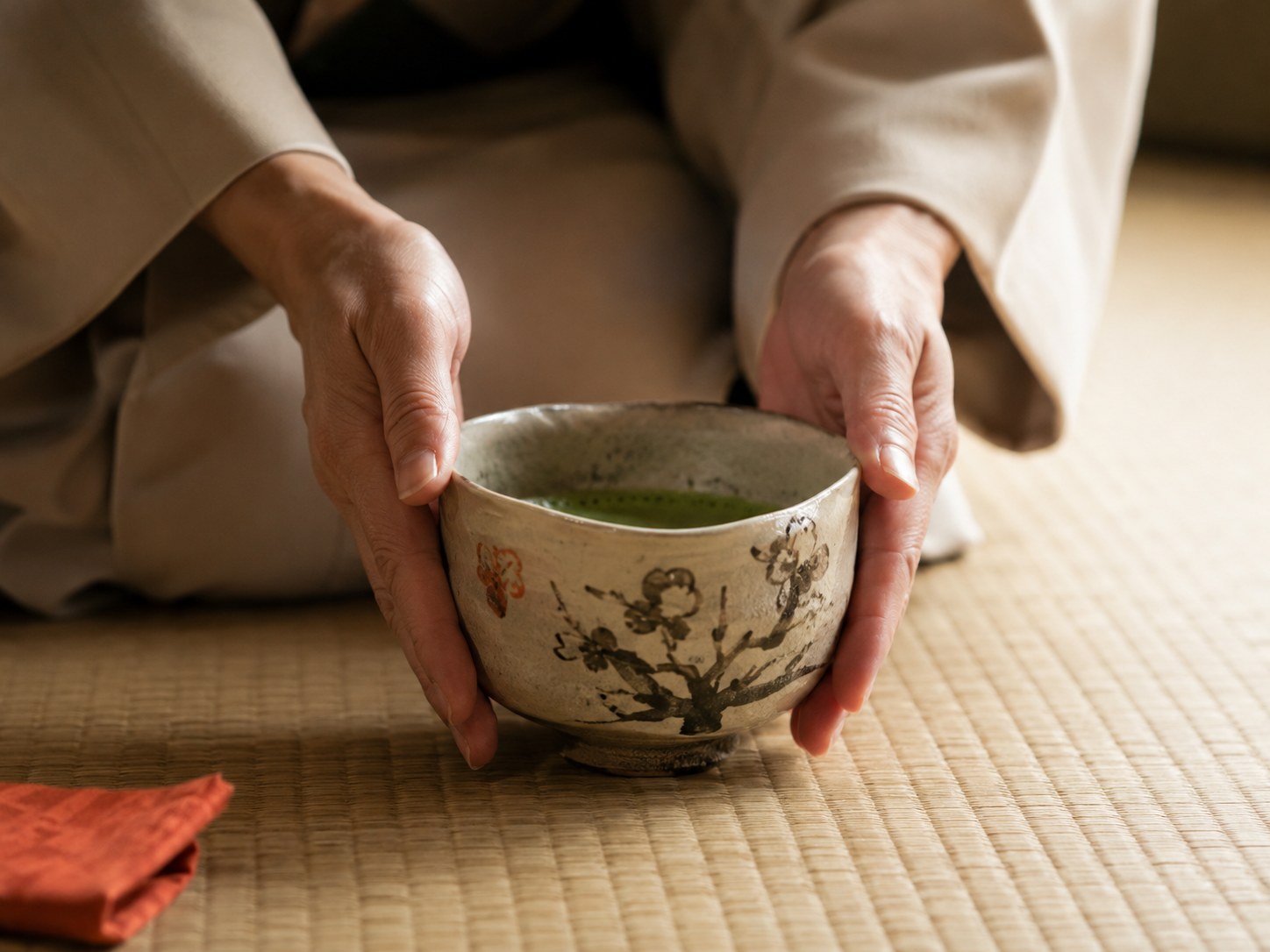 A host's hands placing a matcha tea bowl on the tatami and turning it so its finest face meets the guest, beside a tea room alcove holding a single calligraphy scroll and one seasonal flower — the anticipatory care of omotenashi.