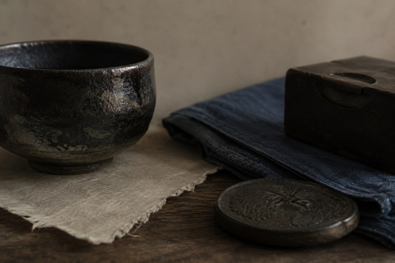 A shibui still life on cream washi: a sober ceramic tea bowl with a deep, muted glaze, a folded length of indigo-dyed cloth, and a dark patinated metal object, all in restrained browns, grays, and indigo — understated, refined, quietly deep.