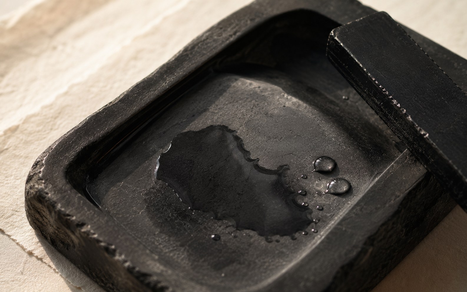 Clear water being added to an inkstone holding a sumi inkstick, droplets catching the light, illustrating that water is one of the two raw materials of ink alongside soot.